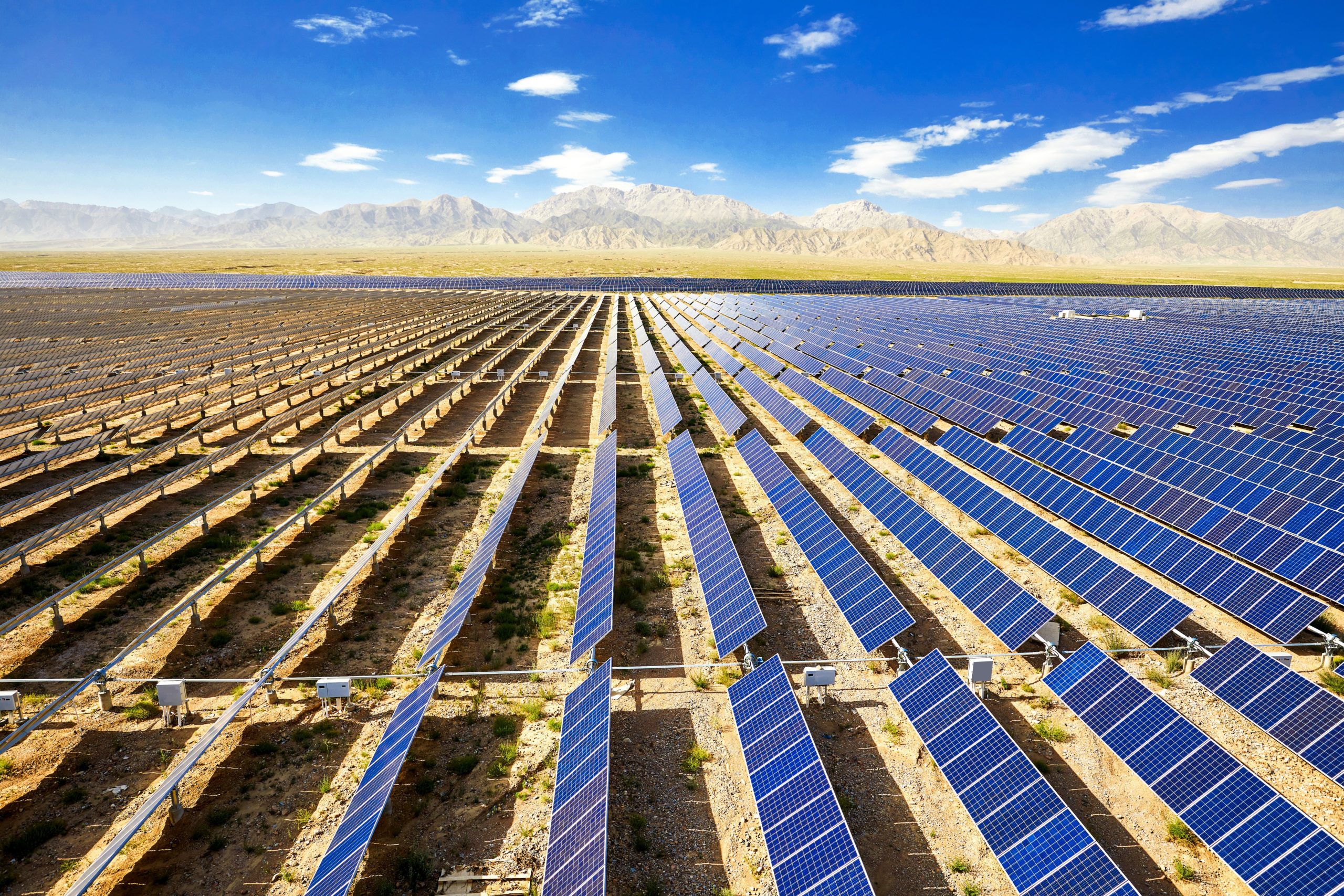 Side view of rows of solar panels in a desert landscape with mountains in the background and a blue sky.