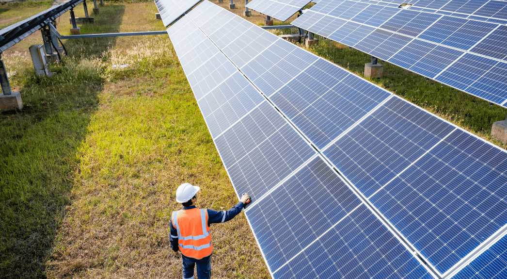 A worker in an orange safety vest and helmet touches a large solar panel in a solar panel farm.