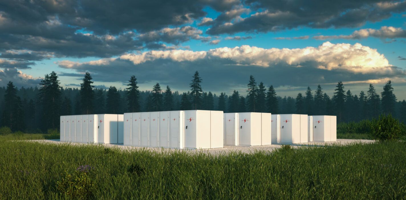 Large white energy storage battery boxes in a grassy field with a forest in the background and a cloudy sky.