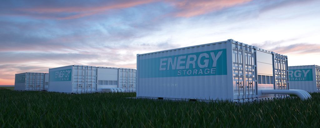 Large, white, battery energy storage boxes in a grassy field with a pink sunset sky.